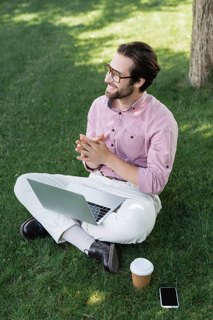 Усміхнений бізнесмен з ноутбуком, дивлячись далеко біля мобільного телефону та кави в парку
  - Фото, зображення