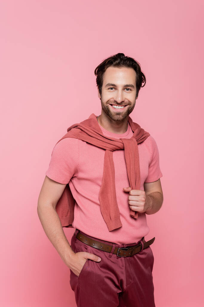 Hombre sonriente sosteniendo la mano en el bolsillo de los pantalones aislados en rosa - Foto, imagen