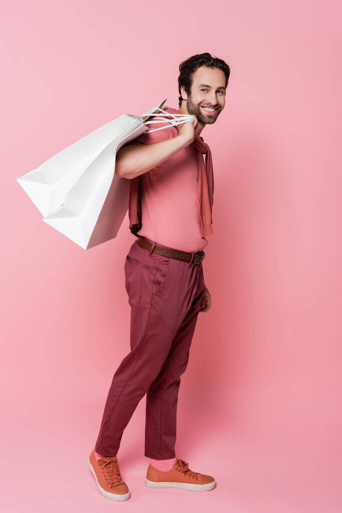 Volle Länge des jungen Mannes mit Einkaufstaschen, der auf rosa Hintergrund in die Kamera blickt - Foto, Bild