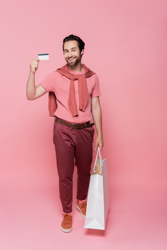 Hombre positivo sosteniendo tarjeta de crédito y bolsa de compras blanca sobre fondo rosa - Foto, Imagen