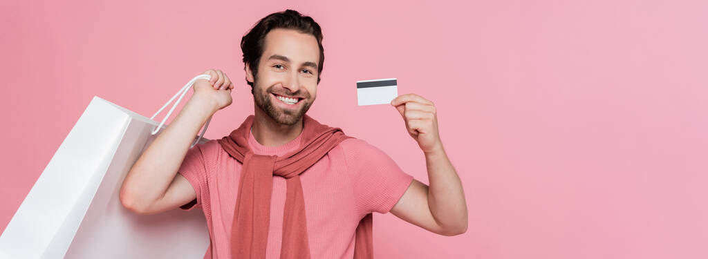 Homme avec sac à provisions montrant carte de crédit isolée sur rose, bannière  - Photo, image