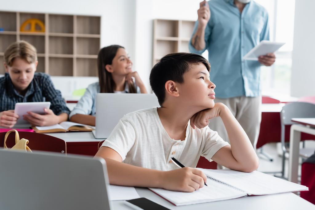 Азіатський школяр, який дивиться у бік, пишучи на записнику біля ноутбука у класі.  - Фото, зображення