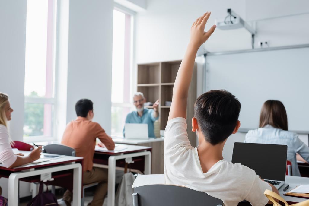 Leerling raisen hand in de buurt laptop en wazig klasgenoten  - Foto, afbeelding