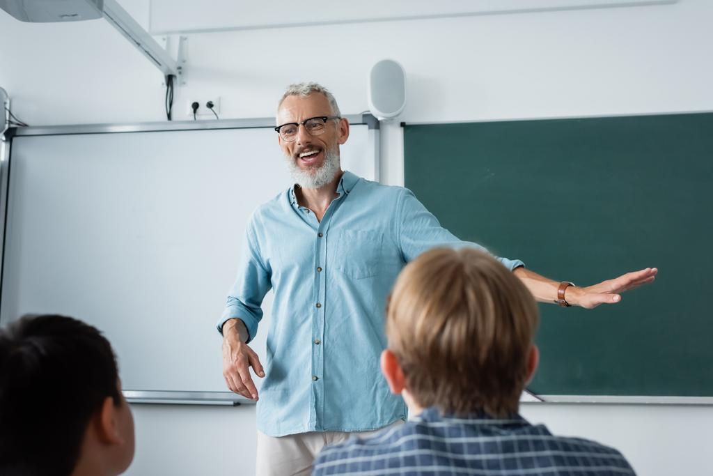 Gülümseyen öğretmen, bulanık okul çocuklarının ve kara tahtanın yanında konuşuyor.  - Fotoğraf, Görsel