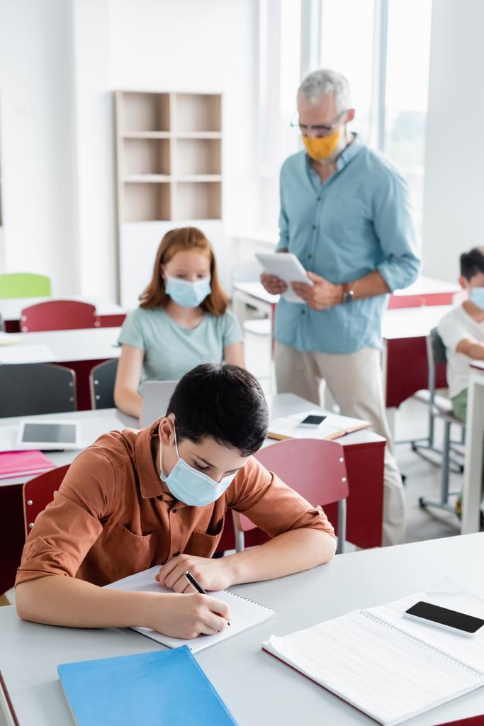 Schulkind in medizinischer Maske schreibt im Klassenzimmer auf Notizbuch  - Foto, Bild