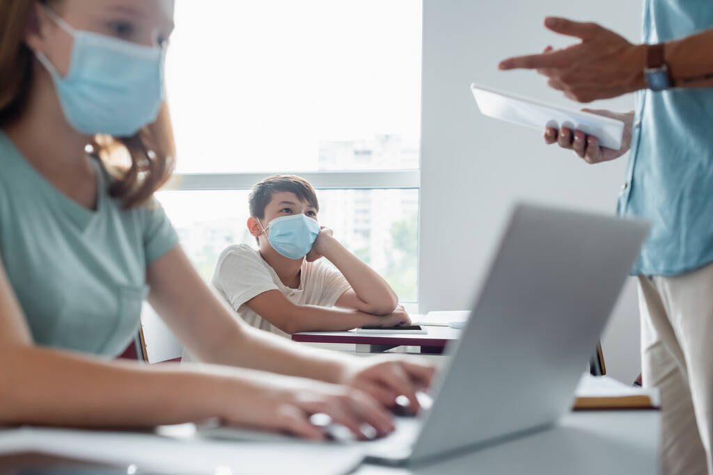 Ασιάτες μαθητές με ιατρική μάσκα κοιτάζοντας θολή δασκάλα με ψηφιακή ταμπλέτα  - Φωτογραφία, εικόνα