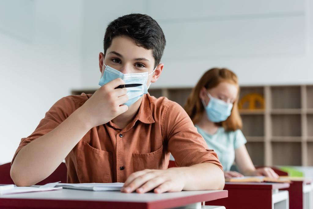 Schooljongen in medisch masker houden pen in de buurt van notebook en wazig vriend  - Foto, afbeelding