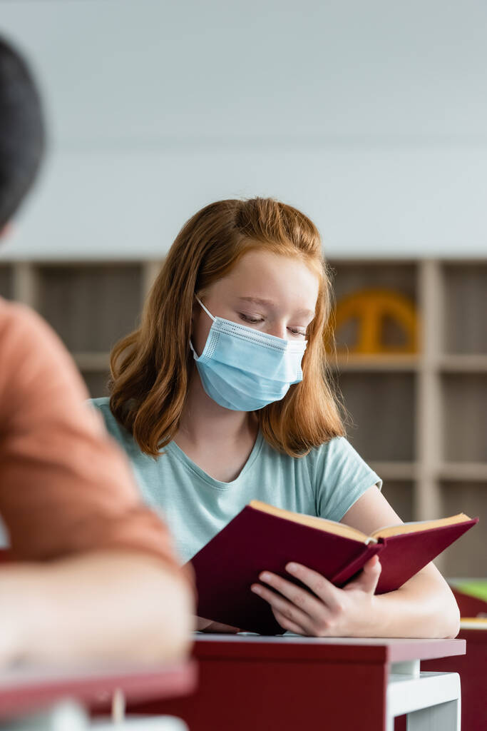 Μαθήτρια με ιατρική μάσκα διαβάζοντας βιβλίο στην τάξη  - Φωτογραφία, εικόνα