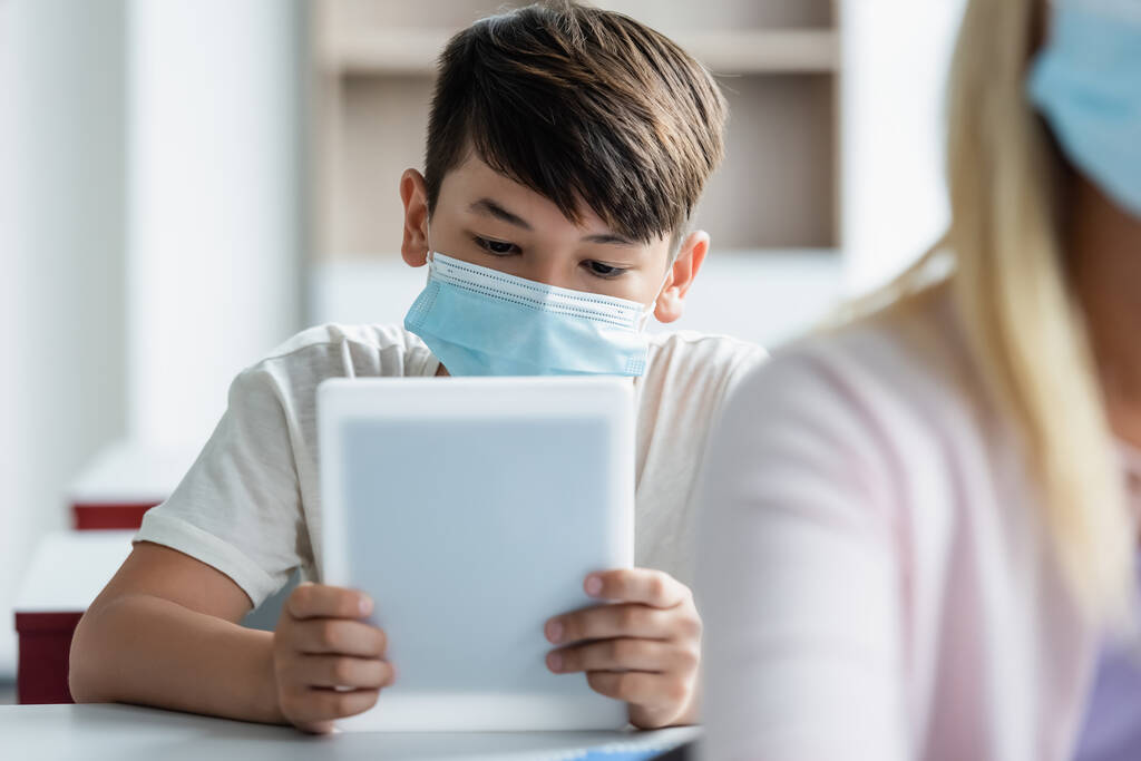 Asiático escolar en médico máscara usando digital tablet en aula  - Foto, imagen