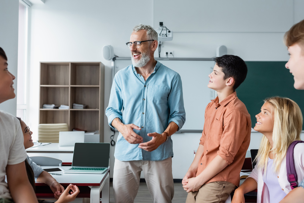 Fröhlicher Lehrer im Gespräch mit Schülern im unscharfen Vordergrund im Klassenzimmer  - Foto, Bild