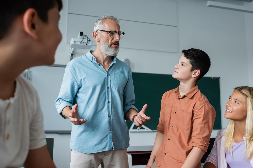 grauhaarige Lehrerin gestikuliert im Gespräch mit Teenagern im Klassenzimmer - Foto, Bild