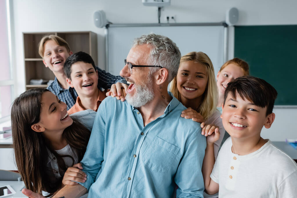 boldog többnemzetiségű tanulók ölelés nevető tanár osztályteremben - Fotó, kép