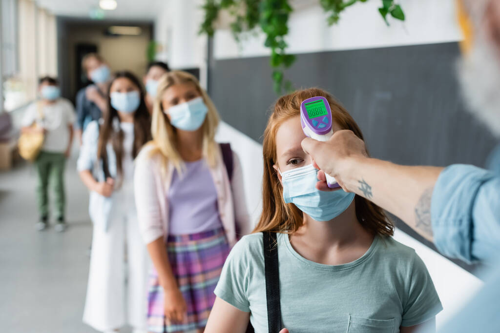 selective focus of pyrometer in hand of teacher checking temperature of pupils standing in line in medical masks - Photo, Image