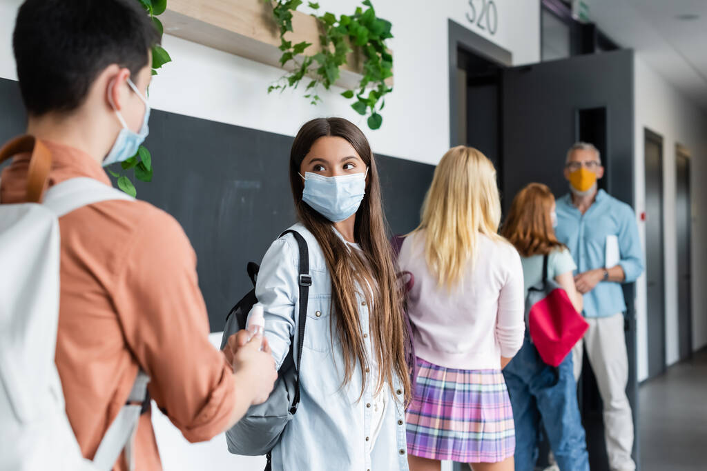 tiener jongen geven hand antiseptisch naar meisje in medisch masker in de buurt wazig klasgenoten en leraar - Foto, afbeelding
