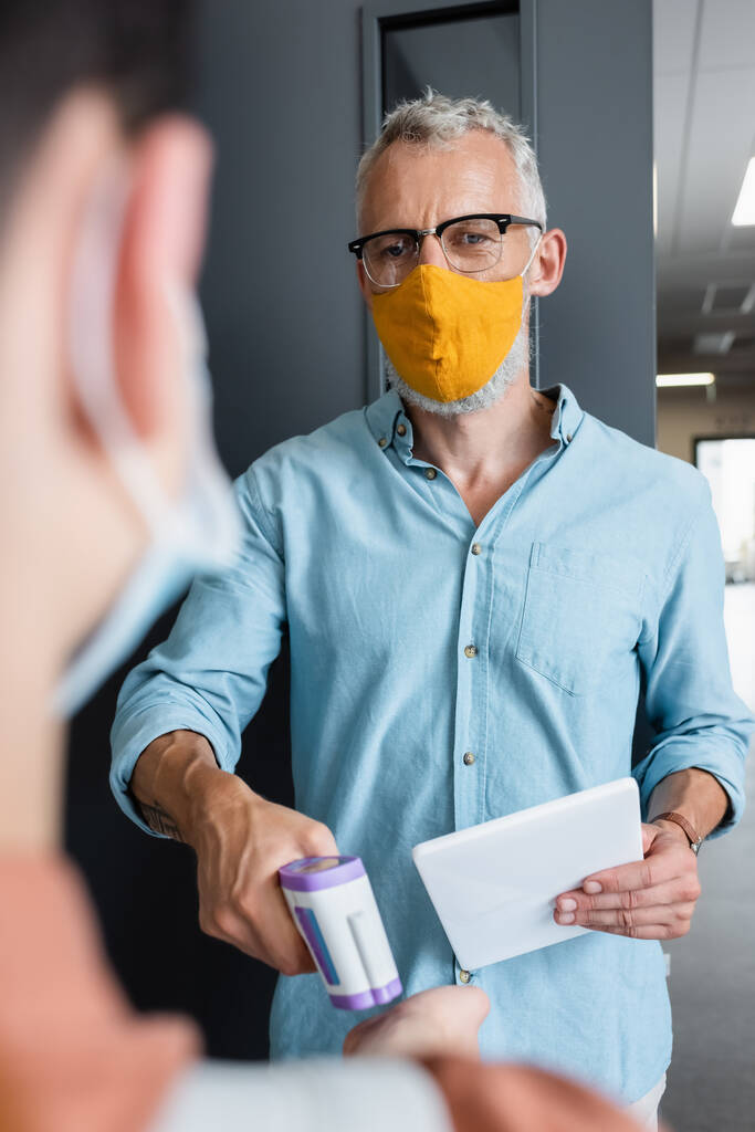 teacher in orange medical mask measuring temperature of blurred boy with pyrometer - Photo, Image