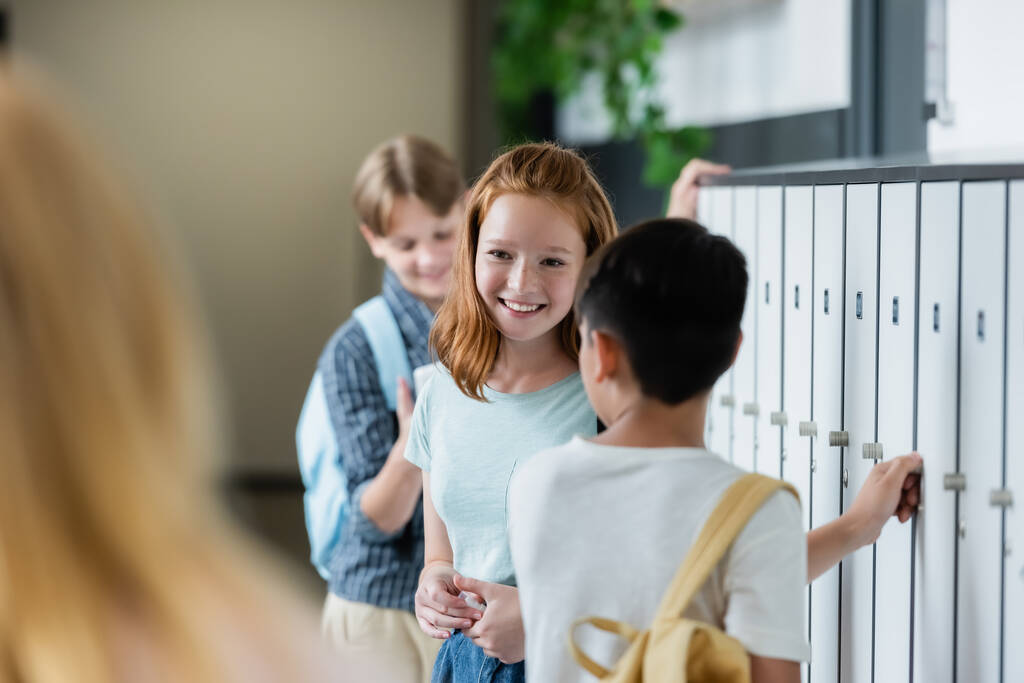 επιλεκτική εστίαση της κοκκινομάλλα μαθήτρια χαμογελώντας στο διάδρομο του σχολείου κοντά θολή έφηβοι  - Φωτογραφία, εικόνα