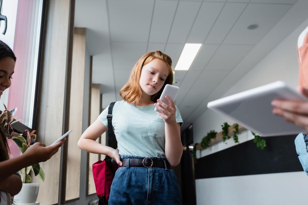 руда дівчина дивиться на мобільний телефон біля розмитих підлітків з гаджетами
 - Фото, зображення