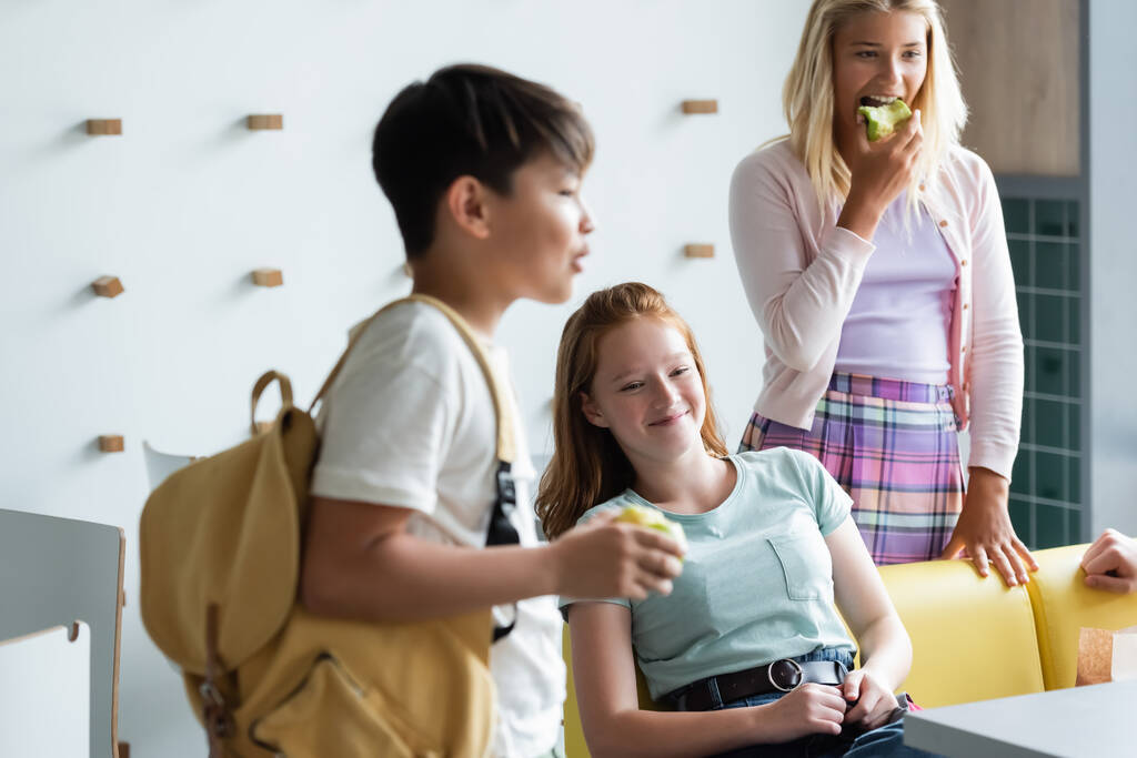 alegre pelirroja chica sentado en la escuela comedor cerca borrosa asiático chico y colegiala comer manzana - Foto, imagen