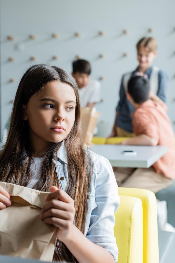 расстроенная девушка сидит с бумажным пакетом в столовой рядом с одноклассниками на размытом фоне - Фото, изображение