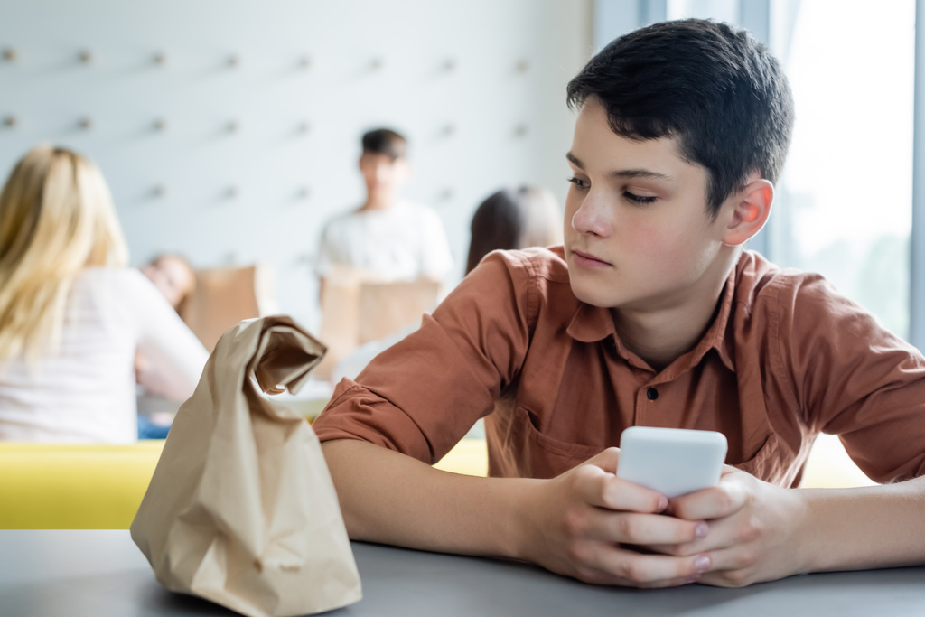 грустный мальчик с мобильным телефоном сидит один в школьной столовой рядом с бумажным пакетом и размытыми одноклассниками - Фото, изображение