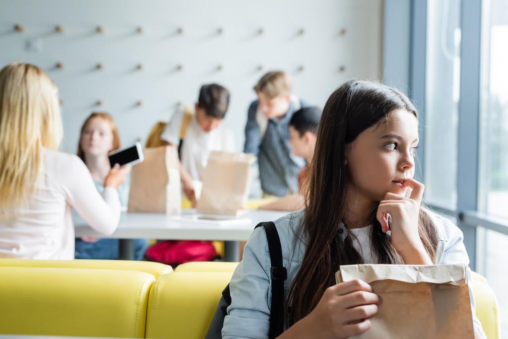 colegiala deprimida mirando hacia otro lado mientras está sentada con una bolsa de papel en la cantina cerca de adolescentes borrosos - Foto, Imagen