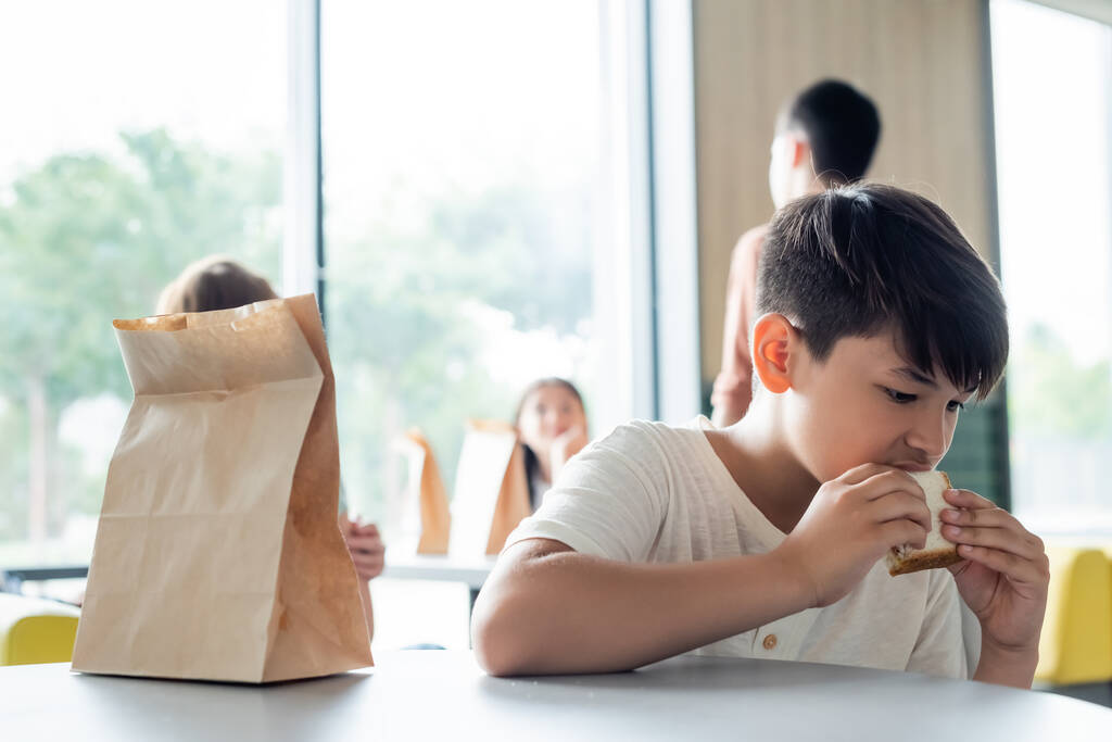 Ασιάτης μαθητής τρώει σάντουιτς μόνος του στην τραπεζαρία κοντά σε θολή εφήβους - Φωτογραφία, εικόνα