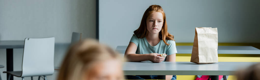 sad girl with sandwich sitting alone in school dining room on blurred foreground, banner - Photo, Image