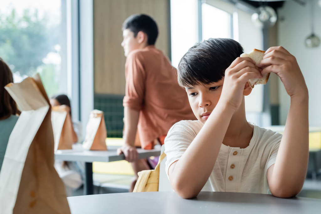 triste asiático chico holding sandwich mientras sentado solo en escuela cantina cerca borrosa pupilas - Foto, Imagen
