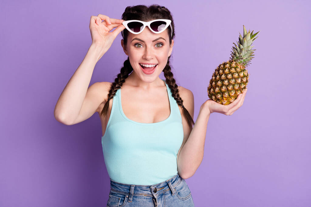 Portret van aantrekkelijke vrolijke meisje aanraken specs houden in de hand sappige ananas geïsoleerd over violet paarse kleur achtergrond - Foto, afbeelding