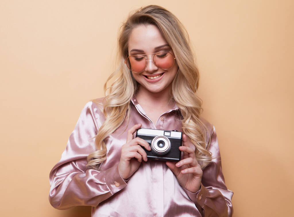 Porträt eines schönen lächelnden jungen Mädchens in rosa Hemd, das isoliert vor beigem Hintergrund steht und eine Fotokamera in der Hand hält. - Foto, Bild