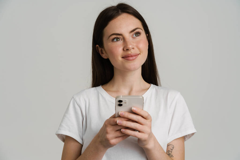 Νεαρή μελαχρινή γυναίκα σε t-shirt χαμογελώντας, ενώ χρησιμοποιώντας το κινητό τηλέφωνο απομονώνονται σε λευκό φόντο - Φωτογραφία, εικόνα