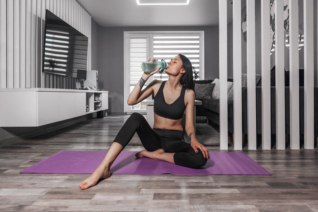 Mulher bonita em sportswear senta-se em um tapete de ioga e bebe água - Foto, Imagem