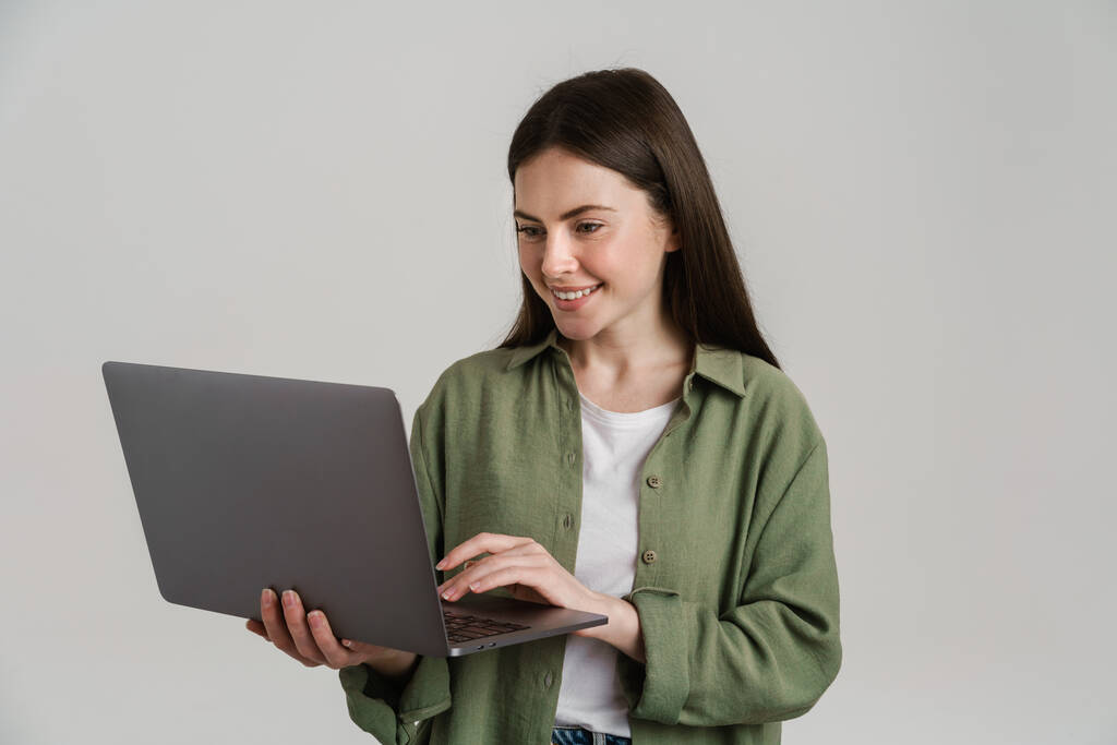 Boldog fiatal fehér alkalmi nő áll gazdaság laptop felett szürke fal háttér gépelés - Fotó, kép