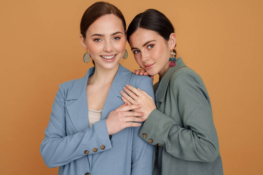 Two smiling elegant women wearing smart jackets posing standing close together isolated over beige background - Photo, Image