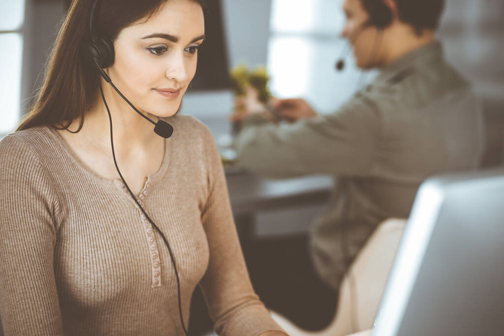 Νεαρό φιλικό κορίτσι σε ακουστικά μιλάει σε έναν πελάτη εταιρειών, ενώ κάθεται στο γραφείο σε ένα σύγχρονο γραφείο μαζί με τη συνάδελφό της. Λειτουργοί τηλεφωνικών κέντρων στην εργασία - Φωτογραφία, εικόνα