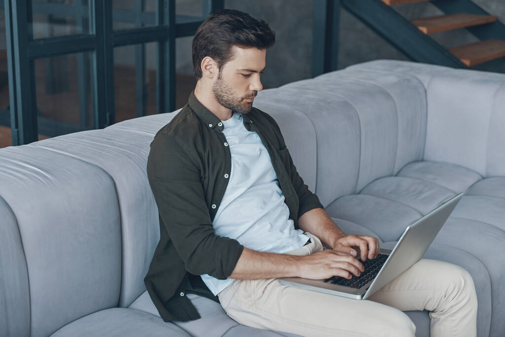 Όμορφος νεαρός άνδρας που χρησιμοποιεί φορητό υπολογιστή, ενώ κάθεται στον καναπέ στο σπίτι - Φωτογραφία, εικόνα