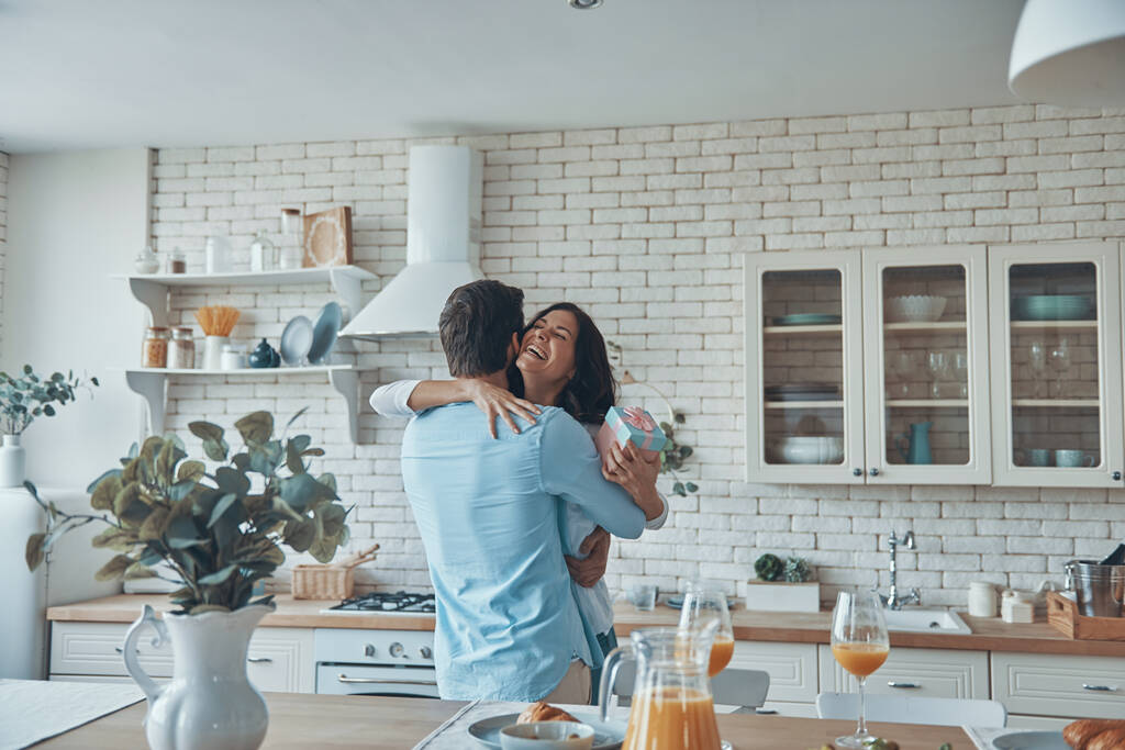 Молодой человек делает подарок своей девушке во время завтрака на домашней кухне вместе - Фото, изображение