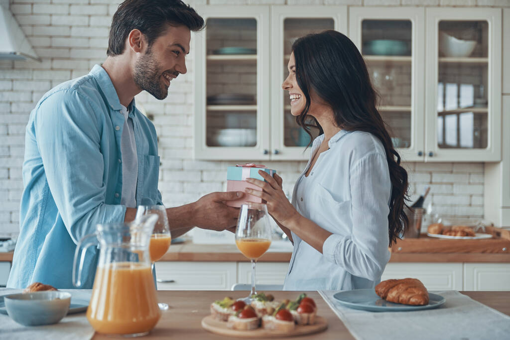 egy fiatalember ajándékdobozt ad a barátnőjének, miközben a konyhában reggelizik. - Fotó, kép