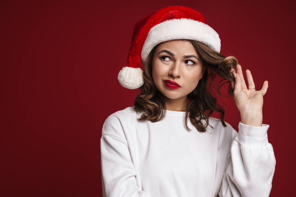 Εικόνα της σκέψης σοβαρή νεαρή γυναίκα στο καπέλο Σάντα Χριστούγεννα απομονώνονται σε φόντο κόκκινο τοίχο - Φωτογραφία, εικόνα