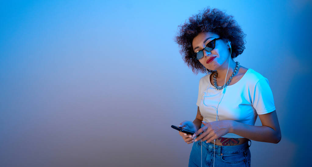 Chica de moda con rizos afro escucha y disfruta de la música con auriculares en luz de neón azul, el estilo hip hop modelo baila y relajarse - Foto, imagen
