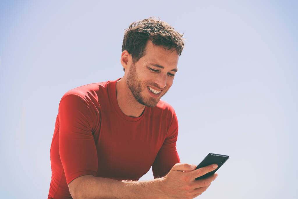 トレーニングセッションの後に携帯電話でビデオを見て健康な男は、モバイルデバイス上のアプリを使用して外に座っている。夏の屋外で彼の20代後半のカジュアルな若い男性大人。青空の背景. - 写真・画像
