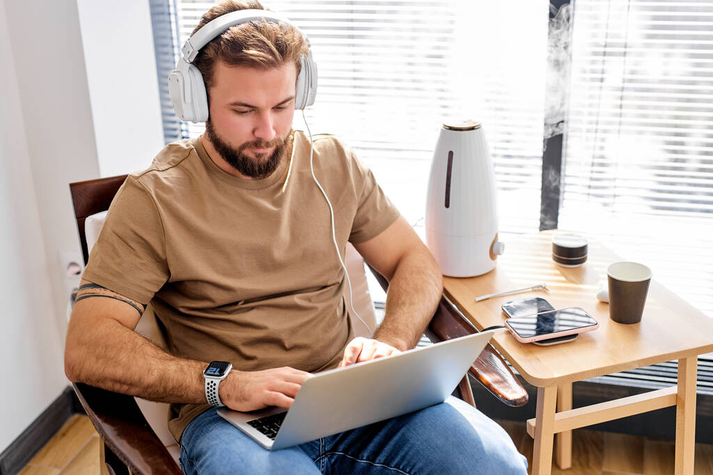 бородатий білий чоловік сидить, працюючи на ноутбуці, з очищувачем в кімнаті, в розумному будинку
 - Фото, зображення