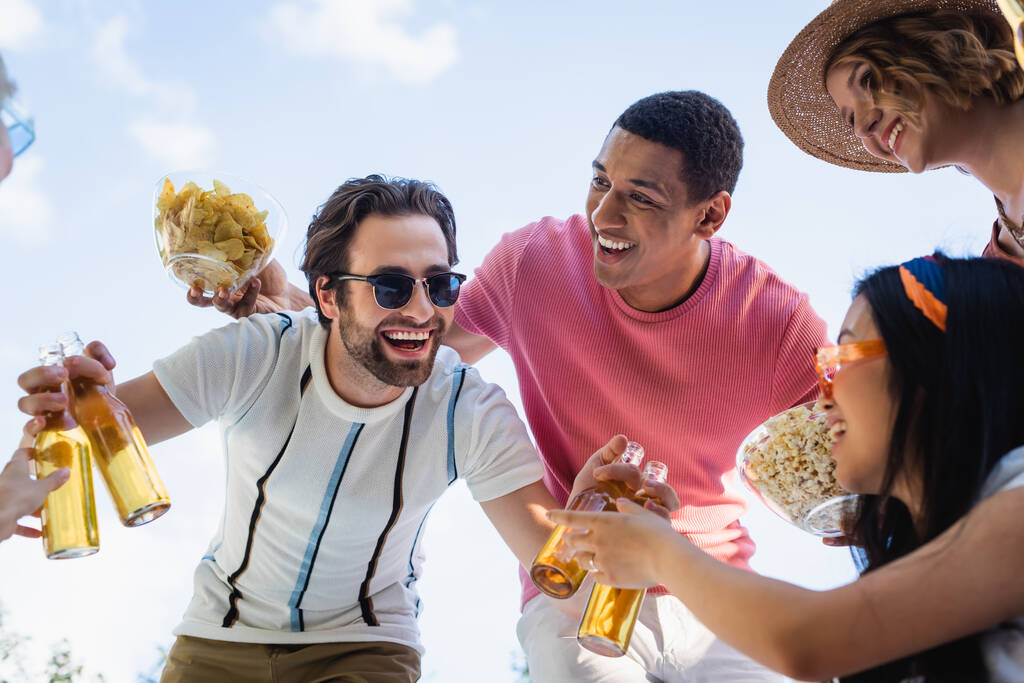 vista de ángulo bajo del hombre alegre en gafas de sol con cerveza cerca de amigos multiétnicos - Foto, imagen