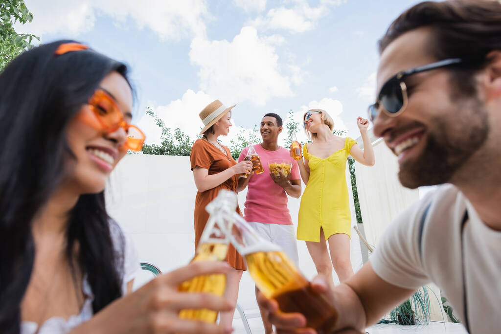 blurred interracial man and woman clinking bottles of beer near happy friends - Photo, Image