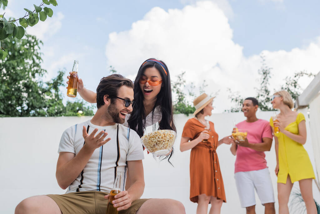 aasialainen nainen ehdottaa popcornia miehelle lähellä monikansallisia ystäviä juomassa olutta hämärtyneellä taustalla - Valokuva, kuva