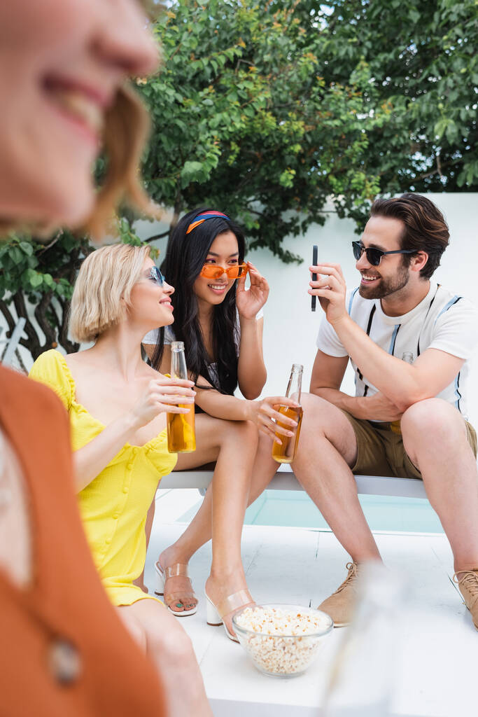 hombre sonriente en gafas de sol mostrando teléfono inteligente a las mujeres interracial durante la fiesta en primer plano borrosa - Foto, imagen
