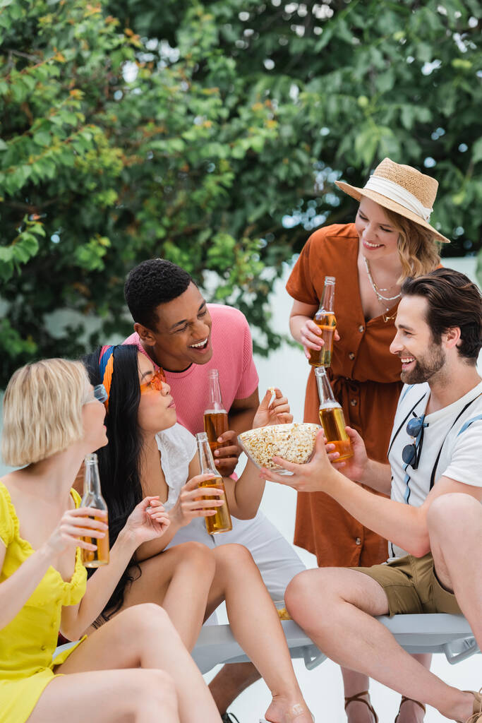 Улыбающийся мужчина держит миску попкорна рядом с веселыми межрасовыми друзьями, пьющими пиво - Фото, изображение