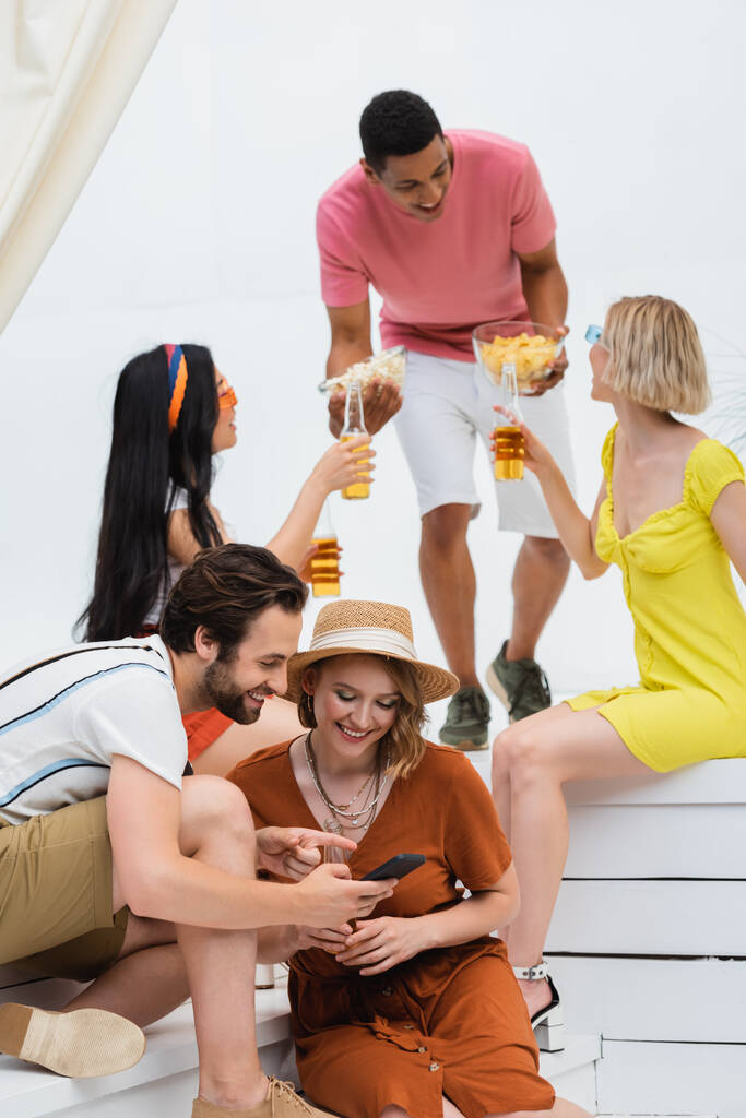 smiling man pointing at smartphone near woman in straw hat while african american man holding snacks near interracial friends - Zdjęcie, obraz
