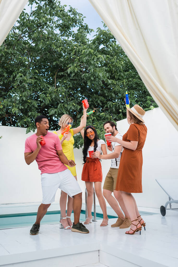 alegres amigos multiétnicos con copas de plástico y pistolas de agua que se divierten en el patio junto a la piscina - Foto, imagen