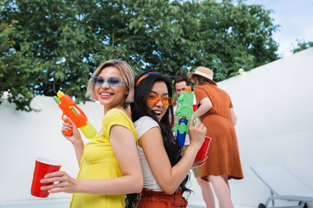 veselé mezirasové ženy s vodní pistole stojící zády k sobě během párty s přáteli - Fotografie, Obrázek